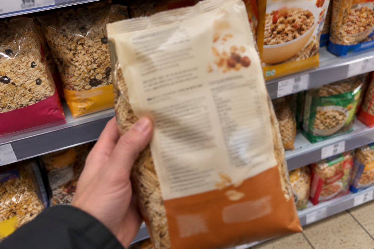 Eine Hand hält die Rückseite eines Getreideprodukts vor einem Supermarktregal; die Beschriftung ist unscharf.